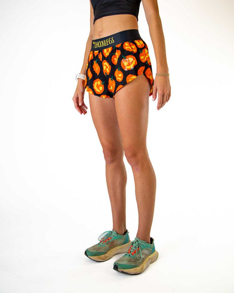 A model wearing black athletic briefs with an orange jack-o'-lantern pattern, paired with green trail running shoes, shown from the waist down on a white studio background.