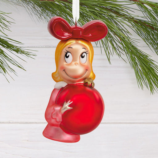 A glossy red Christmas ornament shaped like a smiling girl with blonde hair, a red bow, and a large red gift box, hanging from a white string against a white wood background with green pine branches.