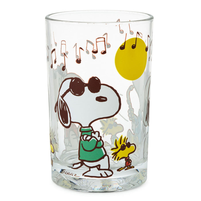 A clear glass tumbler featuring a Peanuts cartoon print of Snoopy wearing sunglasses and a green sweater, with Woodstock, musical notes, a yellow sun, and a gray cloudy background.