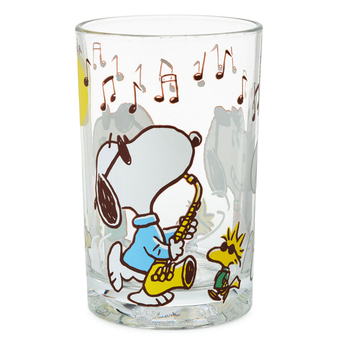 A clear glass drinking tumbler featuring a cartoon print of Snoopy playing a gold saxophone with Woodstock, decorated with brown musical notes against a white background.