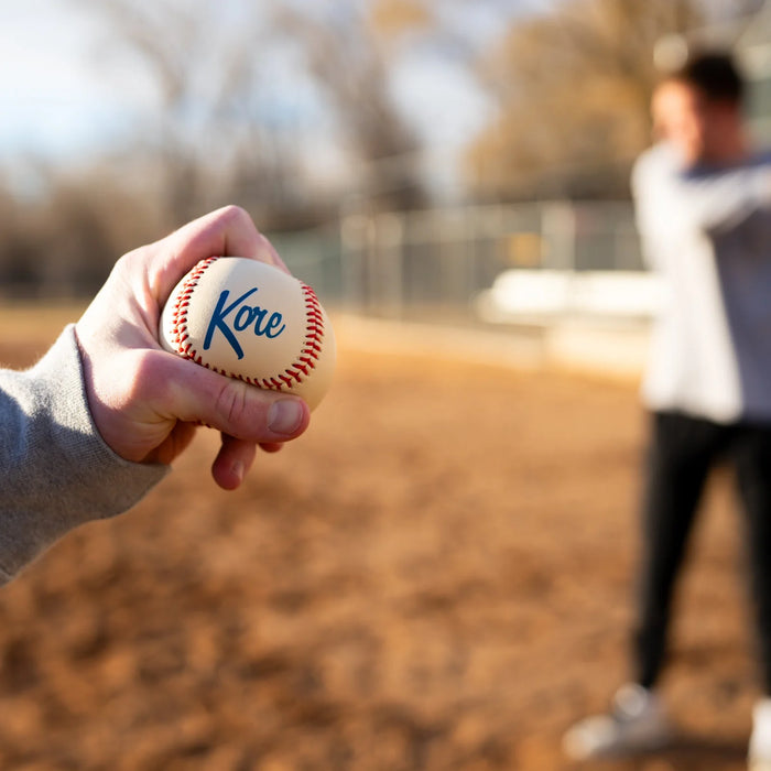 Hand holding a baseball with 'Kore' branding on a blurred outdoor background