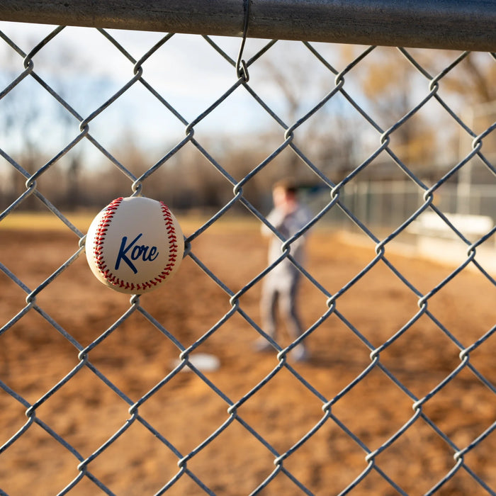 Baseball with 'Kore' branding hanging on a chain-link fence with a baseball player in the background.