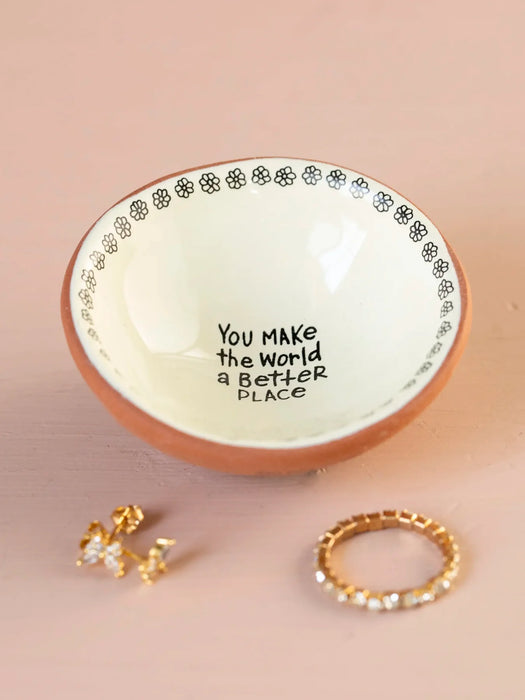 Small ceramic bowl with decorative edge and text, containing gold jewelry on a beige background