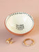 Small ceramic bowl with decorative edge and text, containing gold jewelry on a beige background