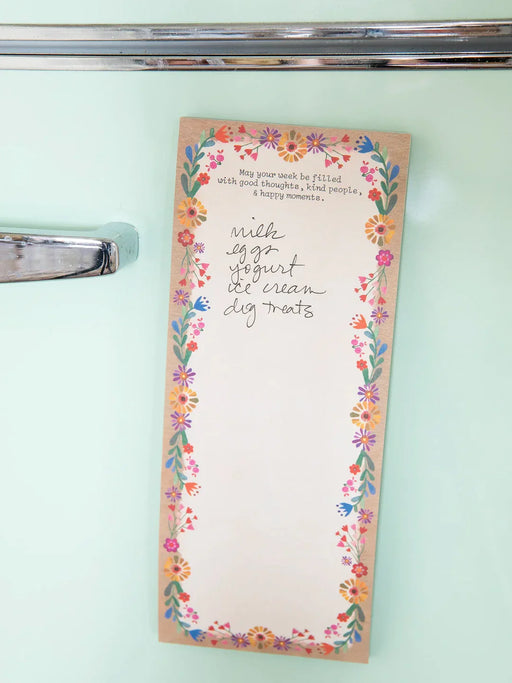 A vertical notepad with a floral border and handwritten text reading 'with egg yogurt ice cream dig treats' is photographed hanging on a mint green refrigerator door next to a chrome handle.