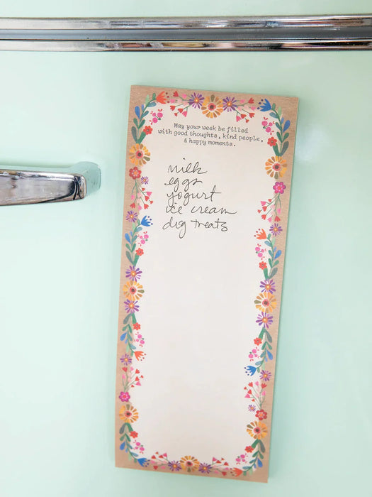 A vertical notepad with a floral border and handwritten text reading 'with egg yogurt ice cream dig treats' is photographed hanging on a mint green refrigerator door next to a chrome handle.