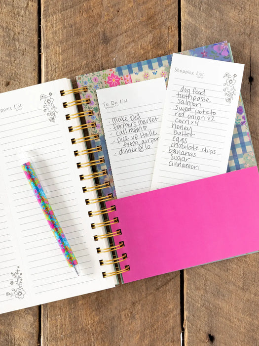 Open planner with various lists and a pink cover on a wooden surface