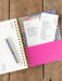 Open planner with various lists and a pink cover on a wooden surface