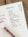 Handwritten shopping list on a notebook with a floral pen.