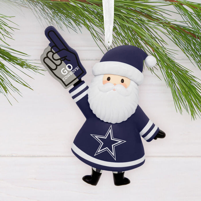 A Dallas Cowboys-themed Santa Claus ornament hanging against a white wood background with pine branches above. The ornament features a navy blue and white Santa figure with a large white beard, a navy hat with a white pom-pom, a white star on the chest, and one hand raised holding a navy foam finger that says 'GO COWBOYS'.