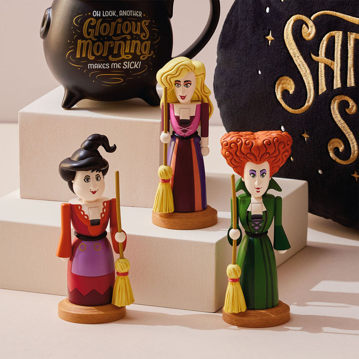 Three witch figurines on wooden bases, styled on white display blocks with a black teapot and a black spherical decor piece in the background.