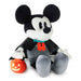 A Mickey Mouse plush toy dressed as a vampire, holding a small orange jack-o'-lantern. The plush features black and white fur-like fabric, gray pants, a black cape, a red bow tie, and a small teal collar accent.