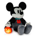 A Mickey Mouse plush toy dressed as a vampire, sitting and holding a glowing jack-o'-lantern pumpkin. The plush features black and white fur, a black cape, a red bow tie, and gray pants, set against a plain white background.
