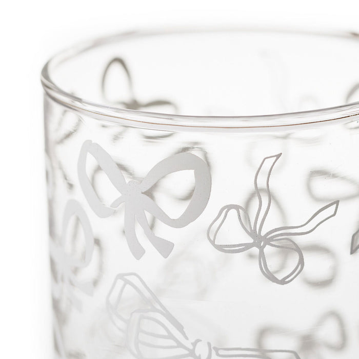 A close-up of a clear glass tumbler featuring a frosted etched pattern of delicate bows in light gray.