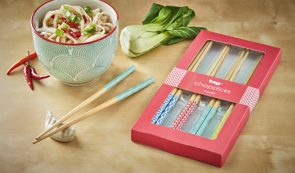 Set of colorful chopsticks with a box on a wooden surface, next to a bowl of noodles and vegetables.