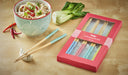 Set of colorful chopsticks with a box on a wooden surface, next to a bowl of noodles and vegetables.
