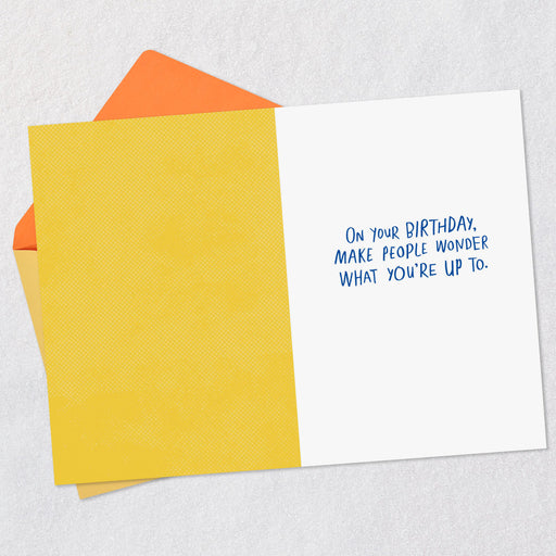 Greeting card with text 'On your birthday, make people wonder what you're up to.' on a yellow card and orange envelope.