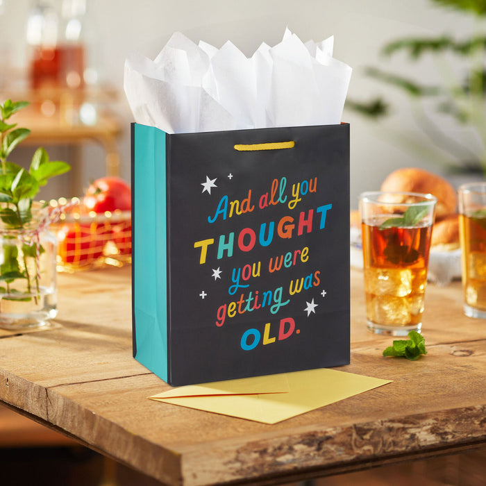 Gift bag with colorful text on a wooden table with drinks and snacks.