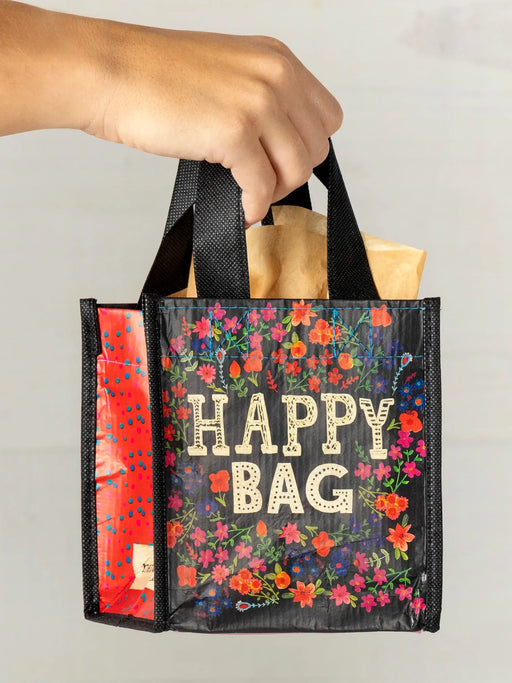 Hand holding a floral 'Happy Bag' with groceries against a neutral background