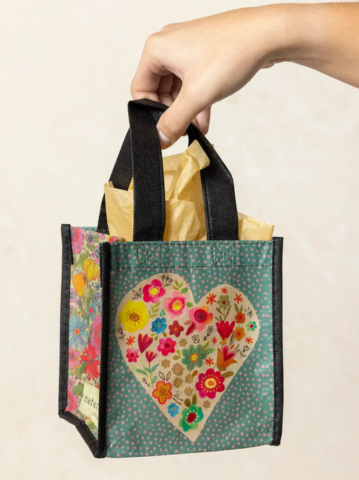 Hand holding a small gift bag with a floral heart design on a beige background