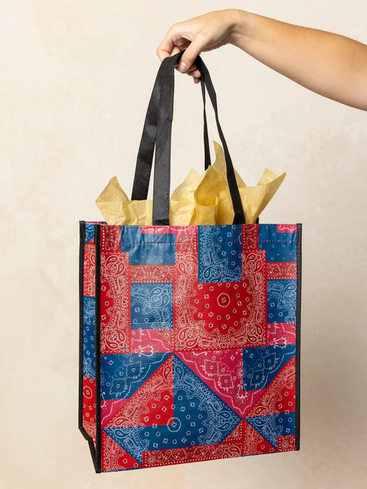 Hand holding a colorful patchwork gift bag with black handles against a beige background