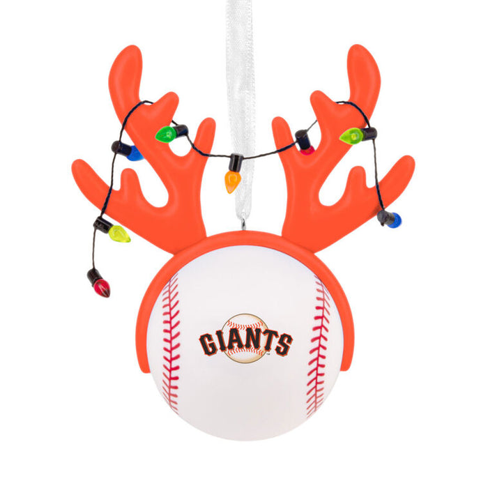 A San Francisco Giants baseball ornament styled as a reindeer antler head, featuring a white baseball with the team logo, orange antlers, and a string of multicolored Christmas lights, hanging from a white ribbon on a white background.