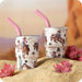 Two tumblers with cowboy designs and pink straws on a sandy surface with flowers.