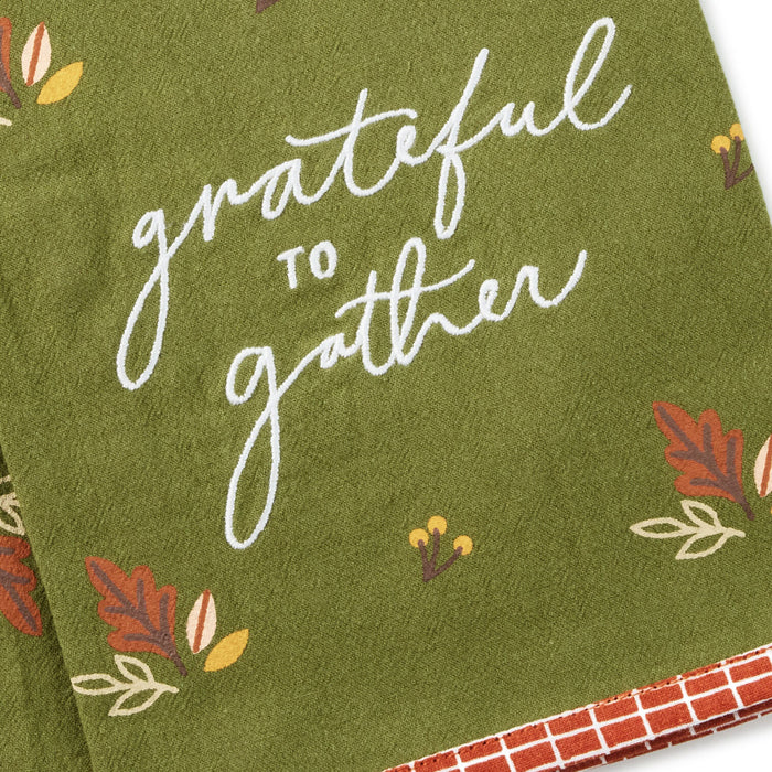 An olive green towel with white cursive text reading 'grateful to gather' and small autumn leaf and berry illustrations, folded with a red-and-white checkered edge visible.