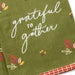 An olive green towel with white cursive text reading 'grateful to gather' and small autumn leaf and berry illustrations, folded with a red-and-white checkered edge visible.