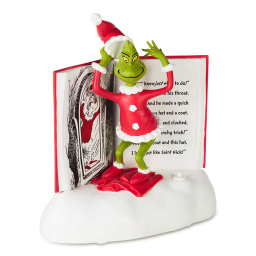 A collectible figurine of the Grinch standing on a white base with an open book behind him, wearing a red Santa hat and coat with white trim and red slippers.