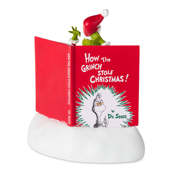 A Dr. Seuss 'How the Grinch Stole Christmas!' book ornament or decorative display featuring an open red book on a white faux-snow base, with a small Grinch figurine peeking out from the top against a plain white background.