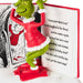 A Grinch figurine in a red Santa-style dress with white trim, posed in front of an open Dr. Seuss book.
