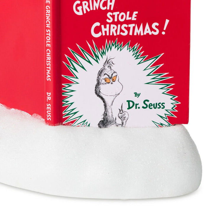 A close-up of a Dr. Seuss 'Grinch Stole Christmas!' book standing upright on a white faux-snow surface, with the red cover and spine visible against a white background.