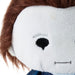 Close-up of a plush doll's face with white fabric, brown hair, a black embroidered eye, and a blue neck area on a white background.