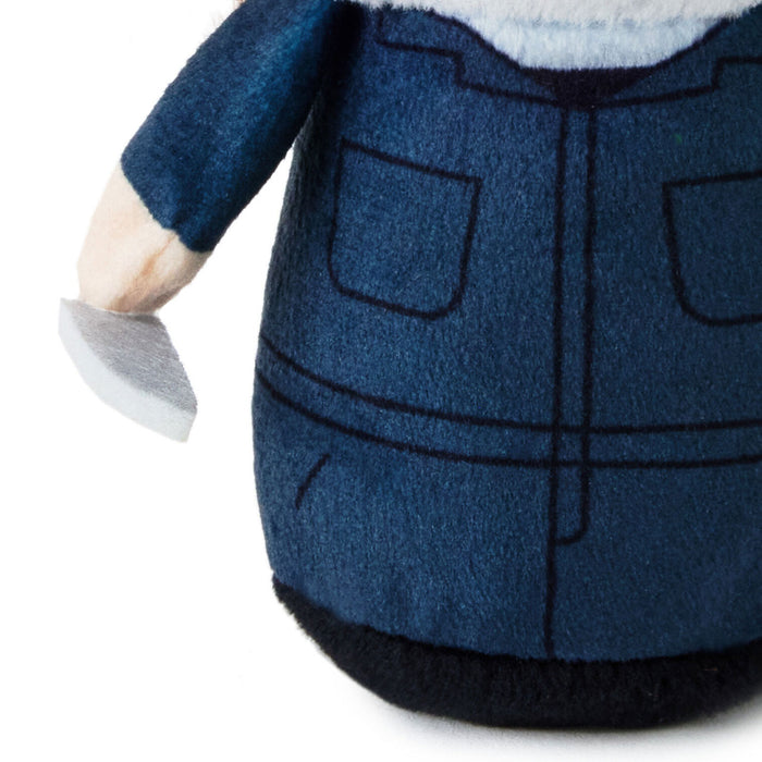 Close-up of a plush toy wearing blue denim-style overalls with black stitched details, a white collar, and a black base, shown against a white background.