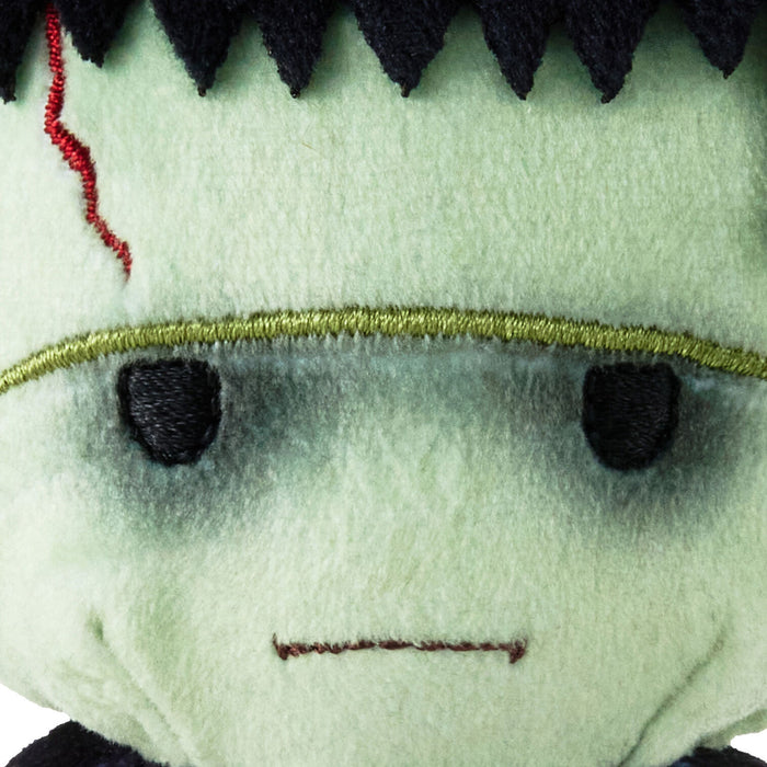Close-up of a plush toy with pale green fabric, black stitched hair, black eyes, a red stitched scar, and a thin gold horizontal line across the forehead.