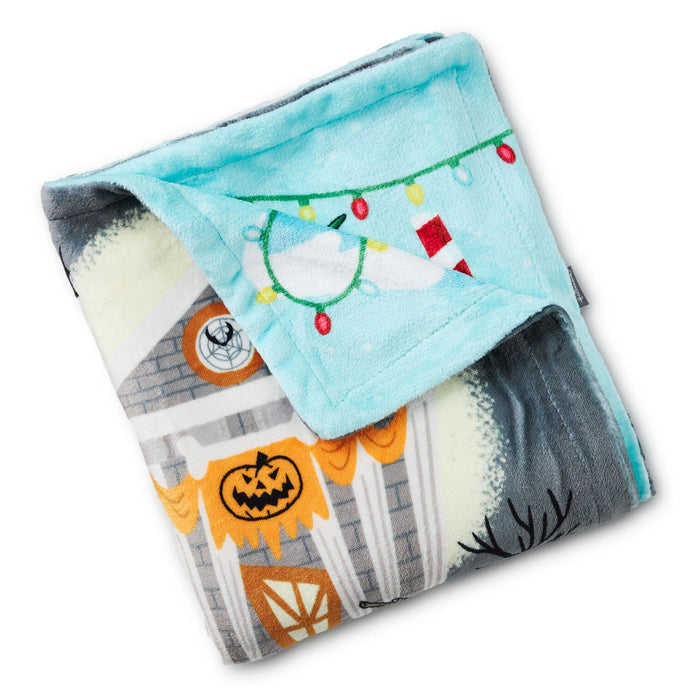 A folded plush blanket with a light blue sherpa-style top and a Halloween-themed printed side showing a haunted house, jack-o'-lantern, moon, and colorful string lights.