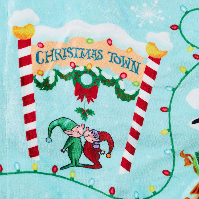 A close-up of a light blue fabric featuring a festive Christmas Town design. The print includes a large banner reading 'CHRISTMAS TOWN' under a snowy archway, decorated with red-and-white striped poles, green holly garlands, colorful Christmas lights, and two kissing elves at the bottom.