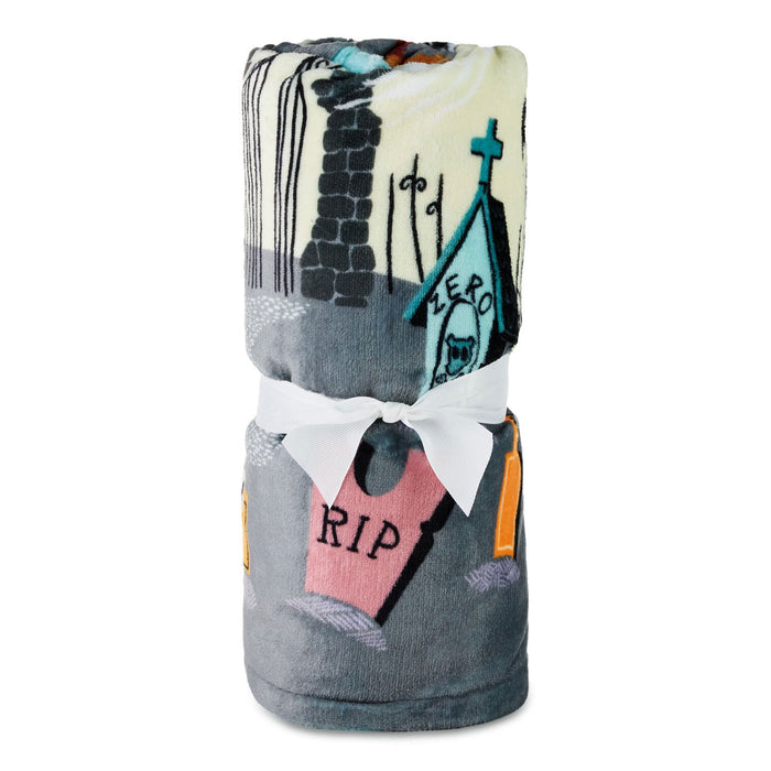 A folded blanket with a Halloween-themed graveyard print, tied with a white ribbon bow. The blanket features a gray base with illustrations of tombstones (one pink with 'RIP' in black), a church steech with a cross, and a cream-colored sky area.