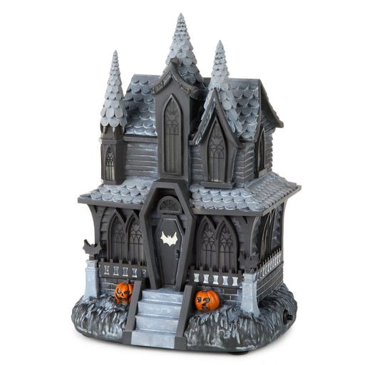 A dark gray gothic haunted house Halloween decoration with pointed spires, a bat-shaped door, small steps, and two orange jack-o'-lantern pumpkins on the base, shown on a plain white background.
