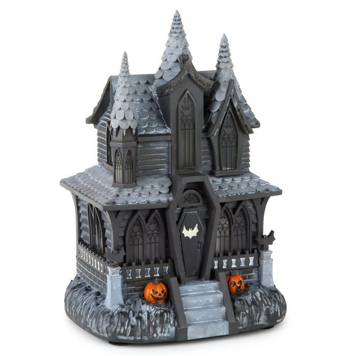 A dark gray plastic haunted house Halloween decoration with pointed spires, arched windows, a bat-shaped door emblem, gray faux-ice detailing on the base, and two small orange jack-o'-lanterns on the front, shown on a plain white background.
