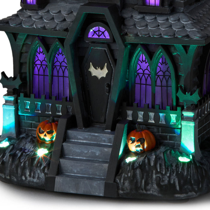 A spooky haunted house Halloween decoration with glowing purple windows, a bat emblem on the door, steps, and two lit jack-o'-lanterns on the front.