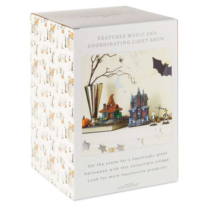 Boxed product packaging for a Halloween-themed collectible village set, shown at a slight angle on a white background. The box is white with a repeating pattern of small haunted houses and features a front photo of the displayed scene with a spooky village, lit bats, and miniature decorations.