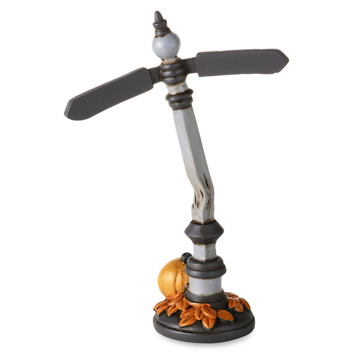 A decorative tabletop clock styled like a vintage directional signpost. It features two dark gray arrow-shaped hands on a weathered silver and black pole, with a small orange pumpkin and autumn leaves at the base, set against a plain white background.