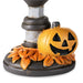 A Halloween-themed decorative base featuring a carved orange jack-o'-lantern pumpkin next to a dark gray and white candlestick, with faux orange leaves around the bottom, shown on a white background.