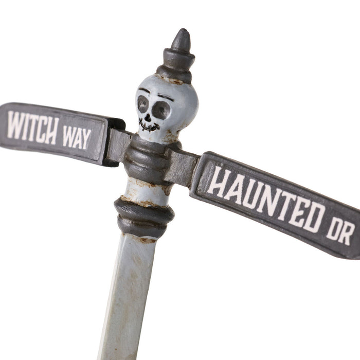 A Halloween street sign decoration featuring a weathered metal T-post with a skull ornament on top, one arm reading 'WITCH WAY' and the other 'HAUNTED DR' in white text on a distressed gray and off-white background.