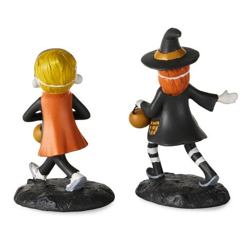 Two small Halloween figurines on a plain white background: one child seen from the back in an orange cape with blond hair, and one witch seen from the side in a black hat and coat holding a pumpkin.