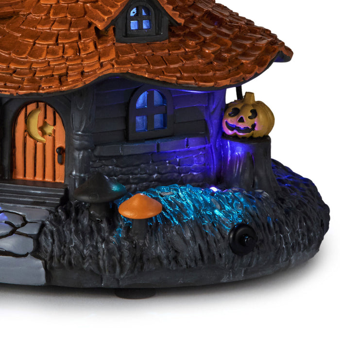 A Halloween-themed miniature house decoration with a brown shingled roof, glowing blue windows, a jack-o'-lantern on the porch, and multicolor LED lighting effects on a white background.
