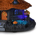 A Halloween-themed miniature house decoration with a brown shingled roof, glowing blue windows, a jack-o'-lantern on the porch, and multicolor LED lighting effects on a white background.