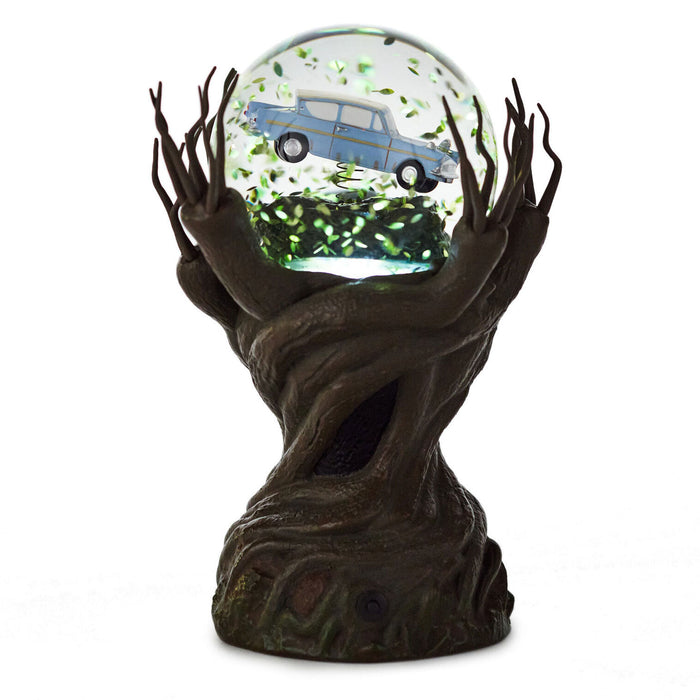 A decorative tabletop sculpture featuring a dark brown tree trunk base with branching arms holding a clear glass globe. Inside the globe is a light blue vintage car surrounded by green leaf-like confetti.